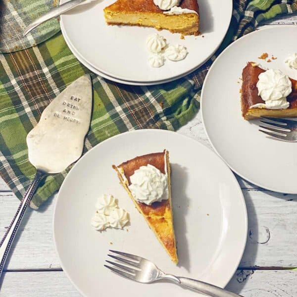 three white plates with pumpkin cheesecake and whipped topping sitting on a green plaid tablecloth with serving spatula