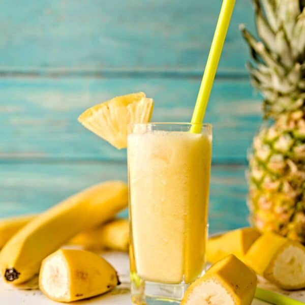 glass full of pineapple smoothie with straw and wedge of pineapple on side and bananas on tabletop