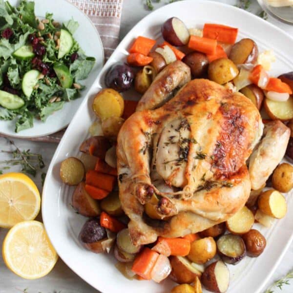 golden roasted chicken and roasted vegetables on white platter with green side salad and yellow lemons on counter