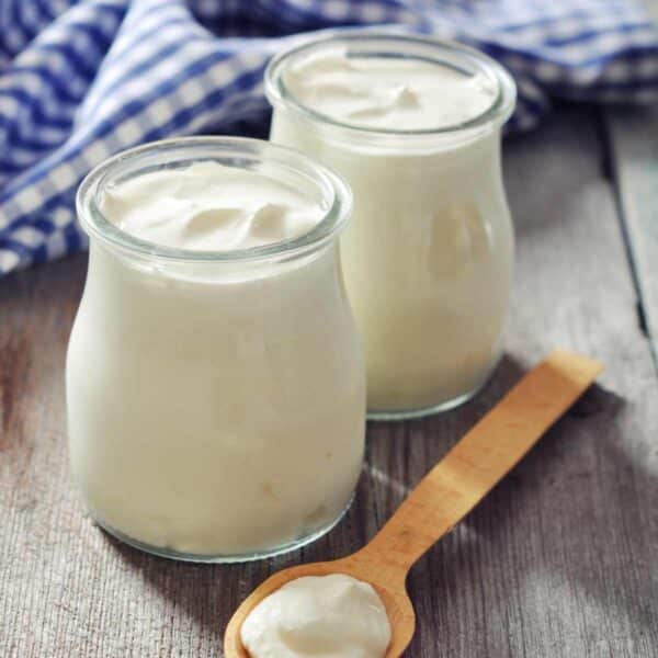 two glass jars of whole milk yogurt on table with wooden spoon and blue gingham towel