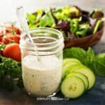 jar of healthy greek yogurt ranch dressing in front of cucumbers, tomatoes, and salad bowl