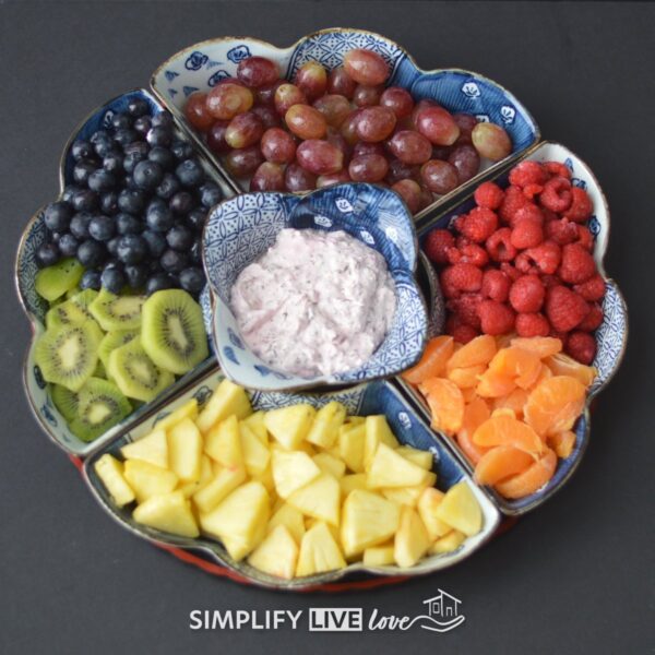 Overhead view of a rainbow fruit tray with raspberries, clementines, pineapple, kiwi, blueberries, and red grapes around a bowl of strawberry yogurt dip