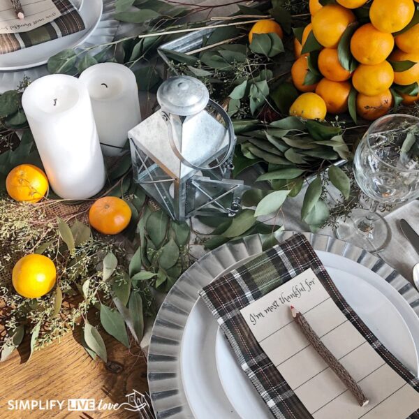 Simple Thanksgiving tablescape with seeded eucalyptus, mandarin oranges, white pillar candles, a galvanized lantern, and a thankful printable on a plaid napkin