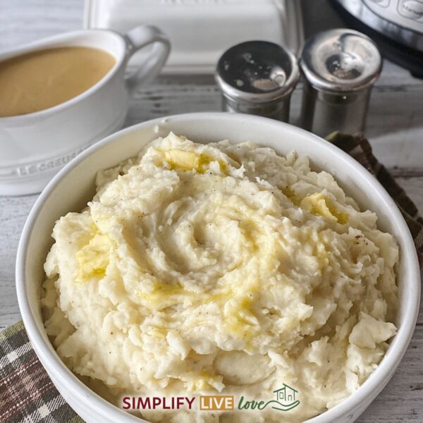 Bowl of creamy Instant Pot mashed potatoes topped with melted butter, with a gravy boat and Instant Pot in the background