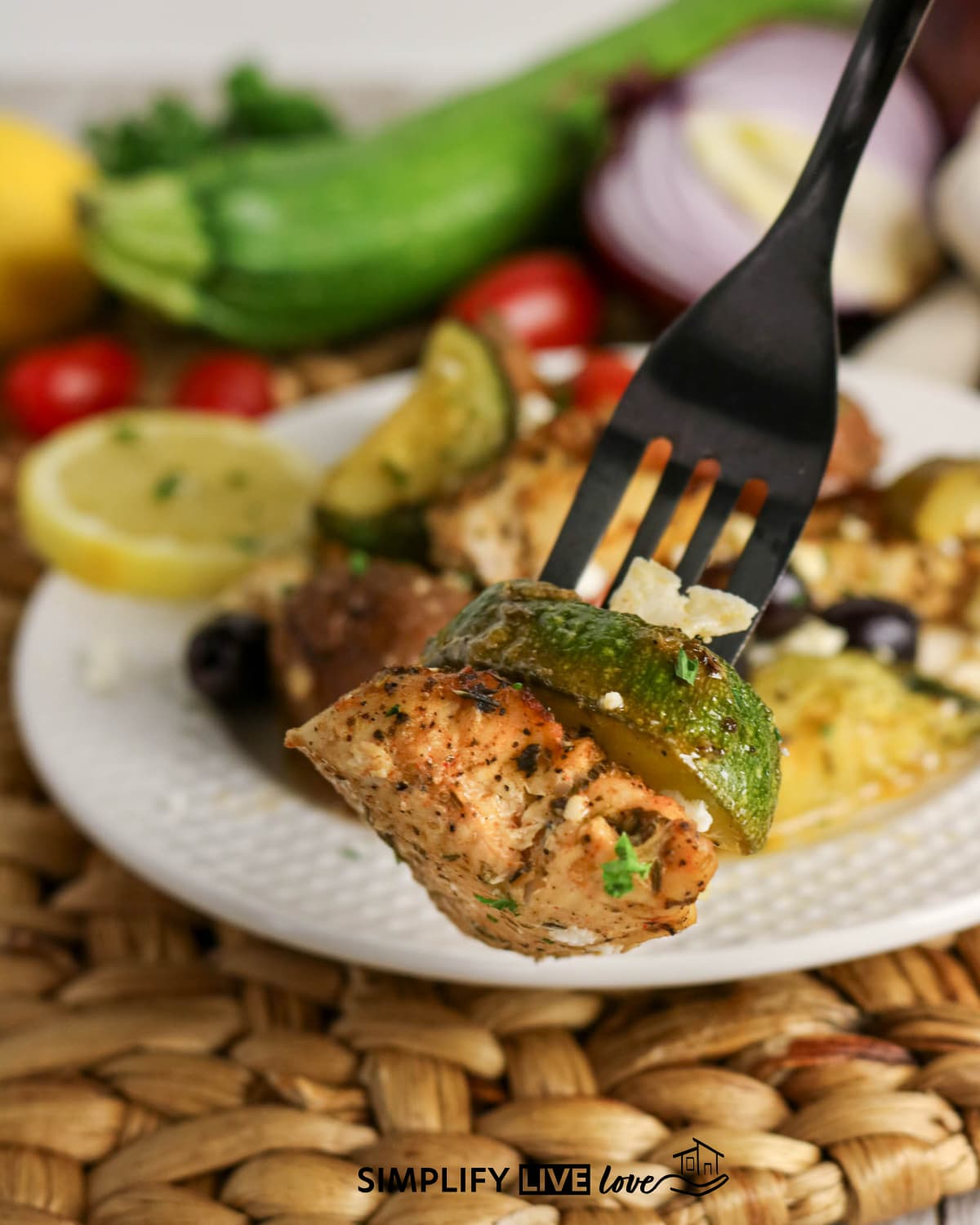 piece of chicken and zucchini on a fork with a plate of greek sheet pan chicken in the background