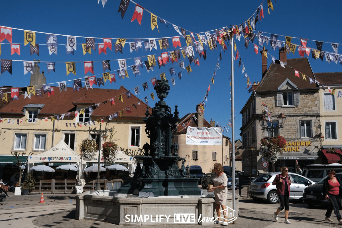 Ornate dark green cast-iron fountain in a town square in Poligny, France, with strings of colorful heraldic bunting overhead, café umbrellas, and flower baskets on stone buildings.