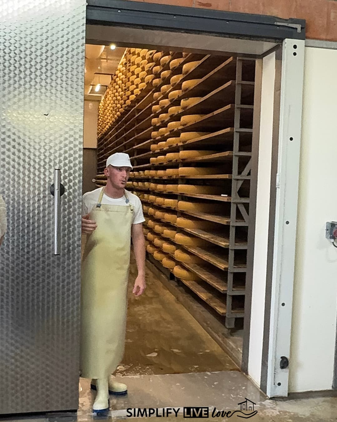 Cheesemaker in an apron standing in the doorway of a dairy cellar with shelves of Comté wheels.