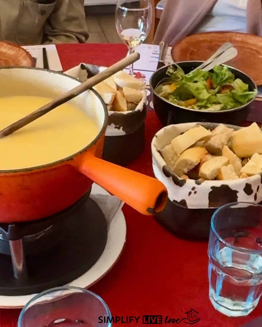 Orange cast-iron pot of melted Comté fondue with bread cubes in cow-print bowls and a green salad on a red tablecloth at Restaurant A la Ferme in Malbuisson.