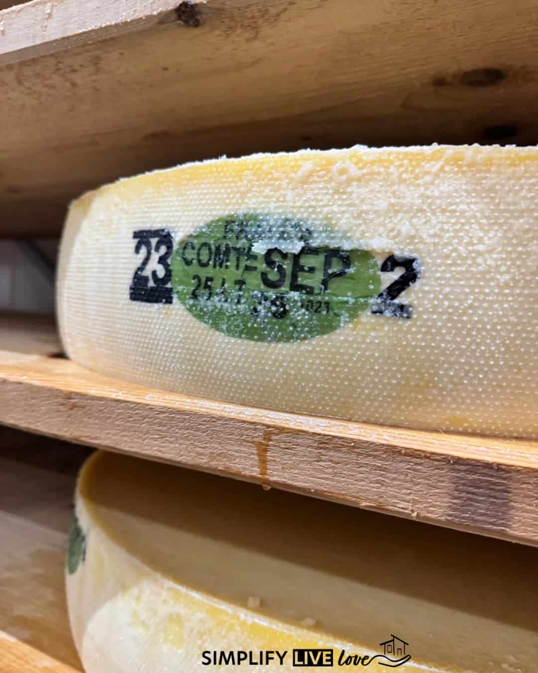 Close-up of a wheel of Comté on a wooden shelf with a green tag pressed into the rind.