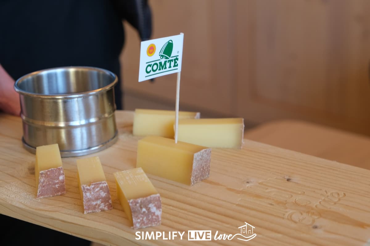 Wooden tasting board with six pieces of Comté cheese, a small metal cup, and a Comté flag stuck in one piece.