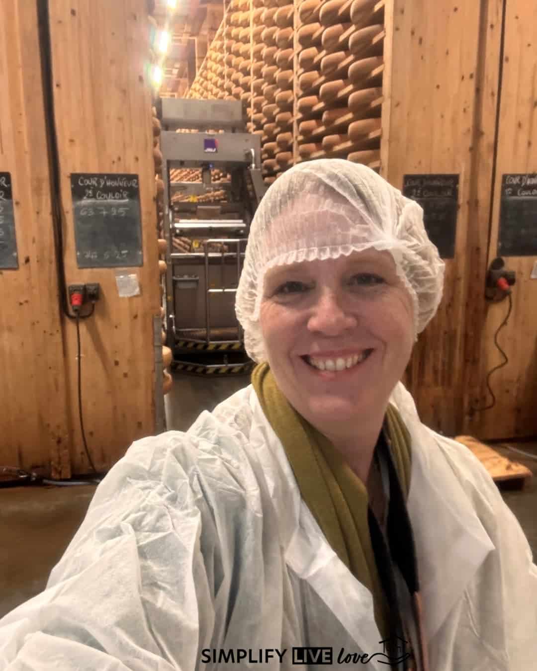 Woman in a hairnet and white coat standing in front of shelves of aging Comté wheels at Fort Saint-Antoine, with a cheese flipping robot in the aisle behind her.