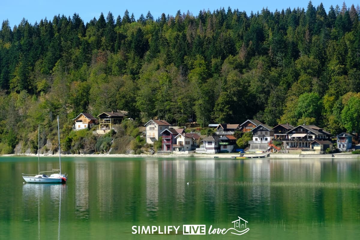 Sailboat moored on the calm green water of Lac de Saint-Point near Malbuisson in the French Jura, with chalet-style houses along the shore below a pine forest.